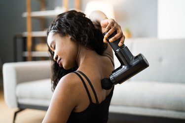 woman using a massage gun on her back and shoulders sitting on the floor at home