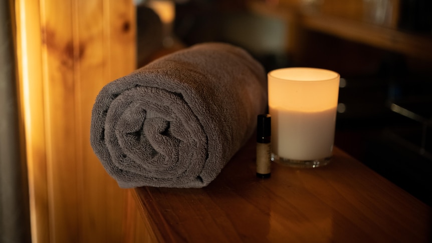 A rolled up grey towel with a lit candle and oil bottle to the right of it sit on a wooden bench in warm lighting.