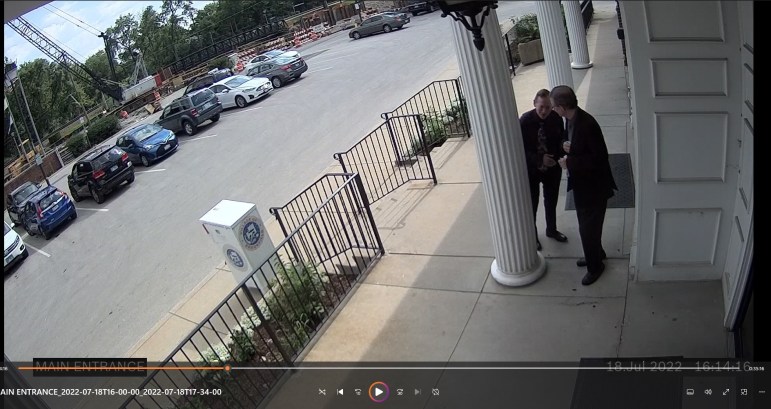 Surveillance video footage of two white men standing next to a white pillar in front of a city hall building. Behind them is a parking lot and construction crane.